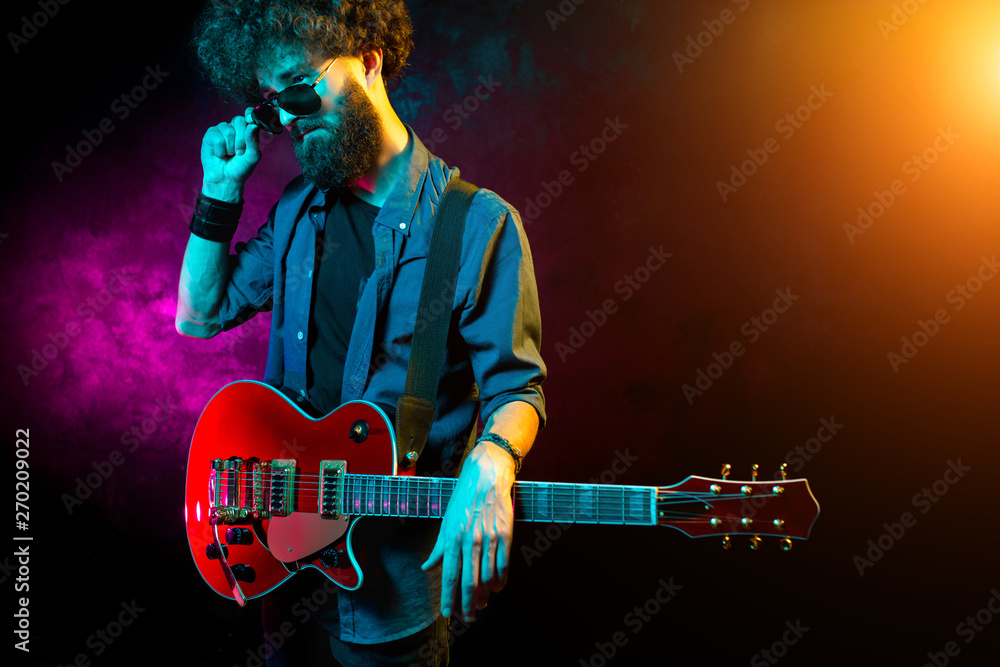 Obraz premium Young hipster man with curly hair with red guitar black sunglasses in neon lights. Rock musician is playing electrical guitar.