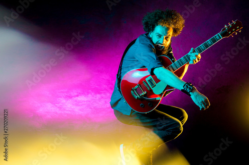 Young hipster man with curly hair jump with red guitar in neon lights. Rock musician is playing electrical guitar.