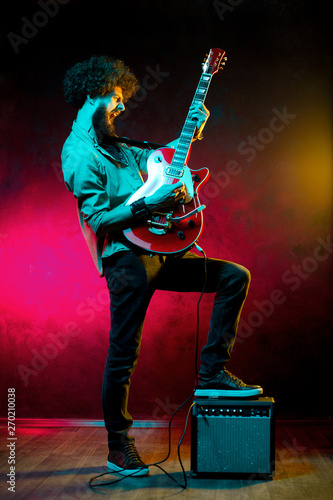 Portrait of hipster man with curly hair with red guitar in neon lights. Rock musician is playing electrical guitar.