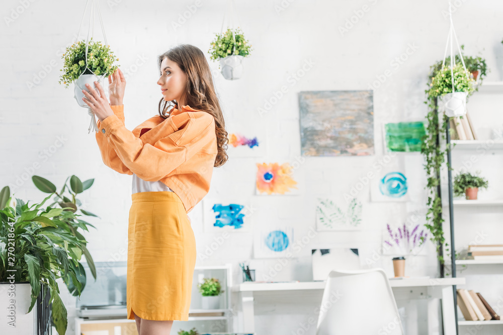 beautiful woman in orange clothing touching hanging flowerpot in spacious room beautiful woman in orange clothing touching hanging flowerpot in spacious room