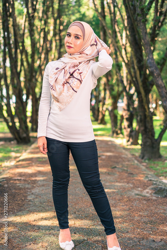 Young attractive Muslim Malay woman, modern contemporary fashion in hijab, close up portrait face and body, blurred background smiling and happy at park.