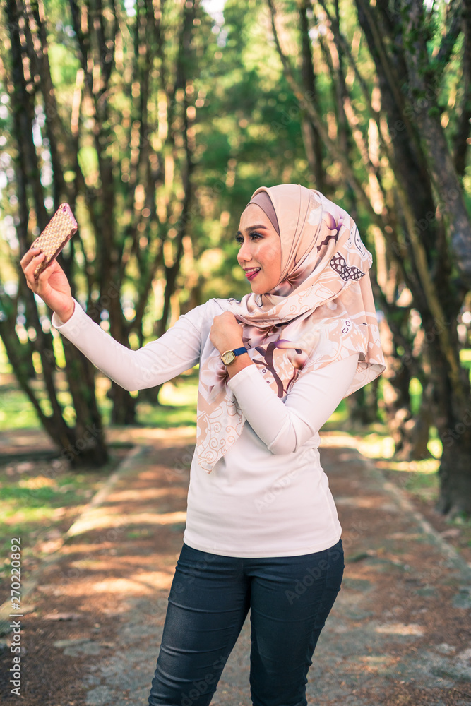 Fashionable beautiful teenage young Muslim Asian girl in hijab and white shirt taking a selfie on smartphone outdoors in park on sunny day. Blurred green nature background copy space. Fashionable beautiful teenage young Muslim Asian girl in hijab and white shirt taking a selfie on smartphone outdoors in park on sunny day. Blurred green nature background copy space.