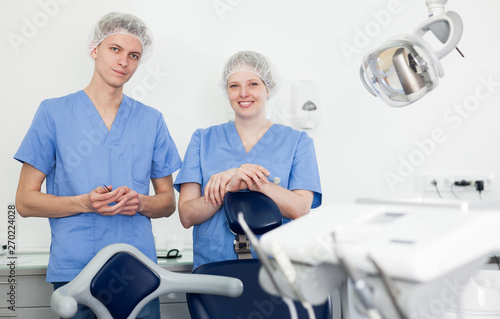 Two professional confident dentists posing near dental chair in clinic