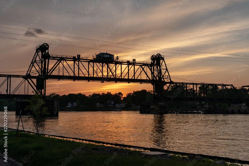A movable, vertical-lift railroad trestle bridge over a river at sunset. Concepts of industry, transportation, urban landscape