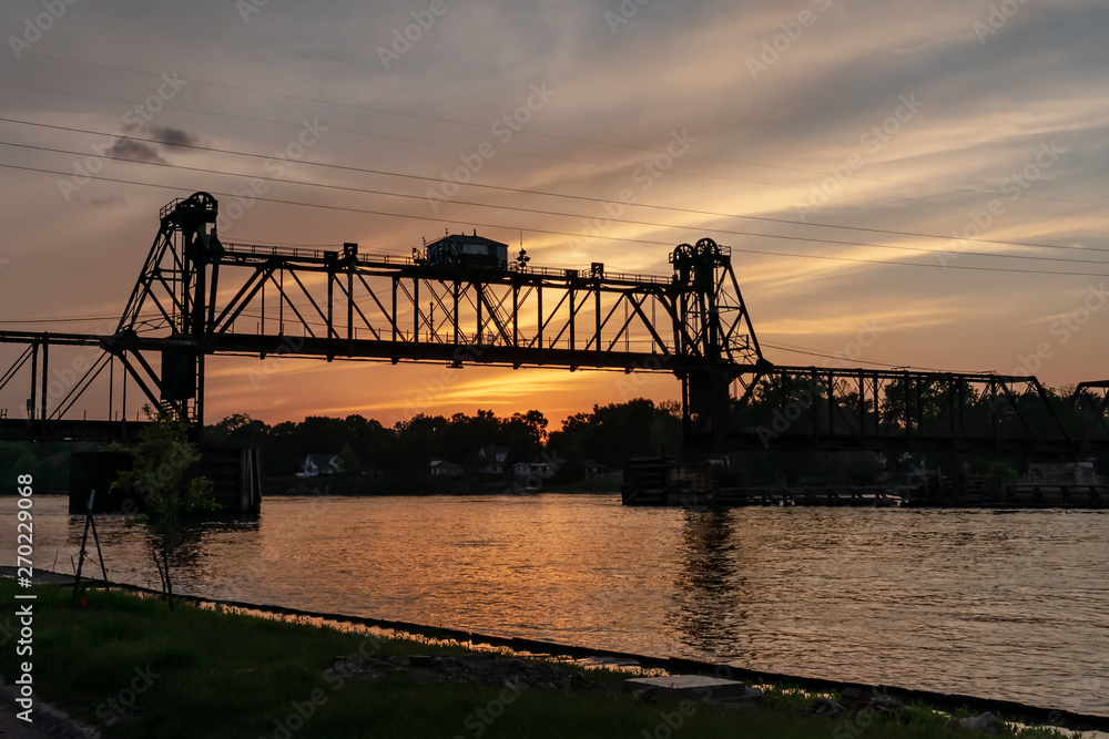 Obraz premium A movable, vertical-lift railroad trestle bridge over a river at sunset. Concepts of industry, transportation, urban landscape