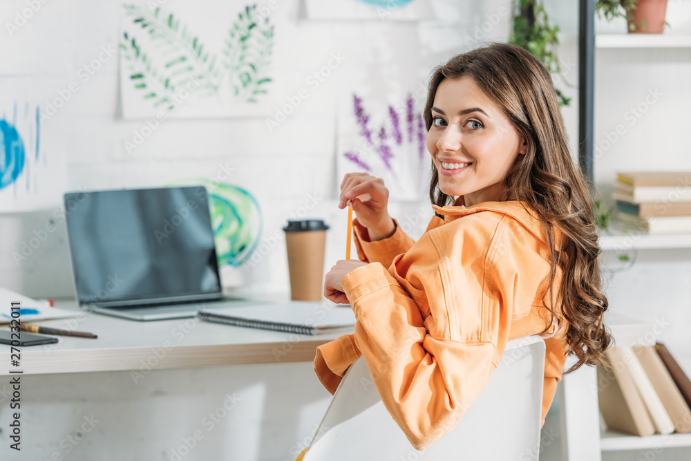 cheerful girl smiling at camera while sitting at desk near laptop with blank screen cheerful girl smiling at camera while sitting at desk near laptop with blank screen