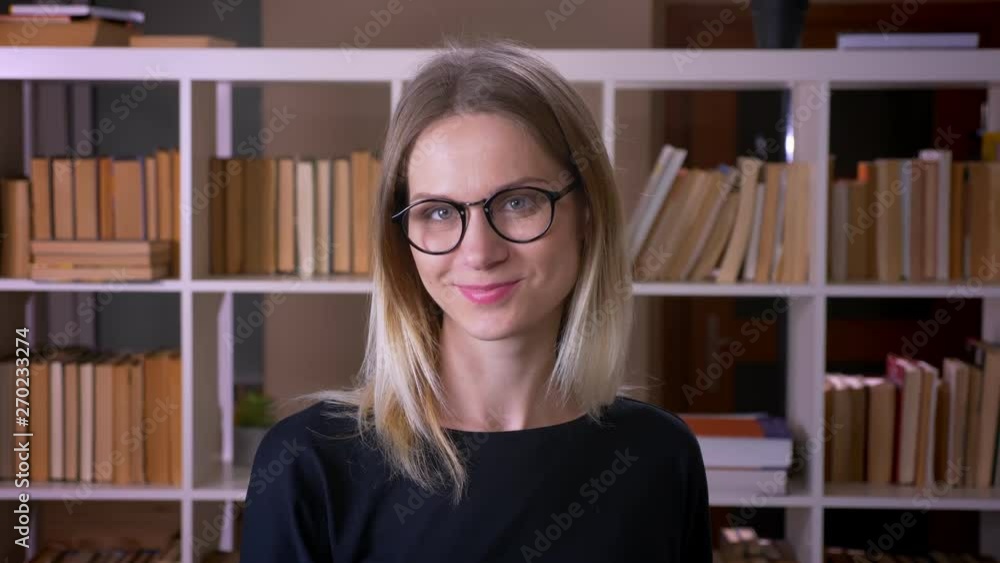 Closeup shoot of young attractive female student in glasses smiling looking at camera indoors in the university library Closeup shoot of young attractive female student in glasses smiling looking at camera indoors in the university library