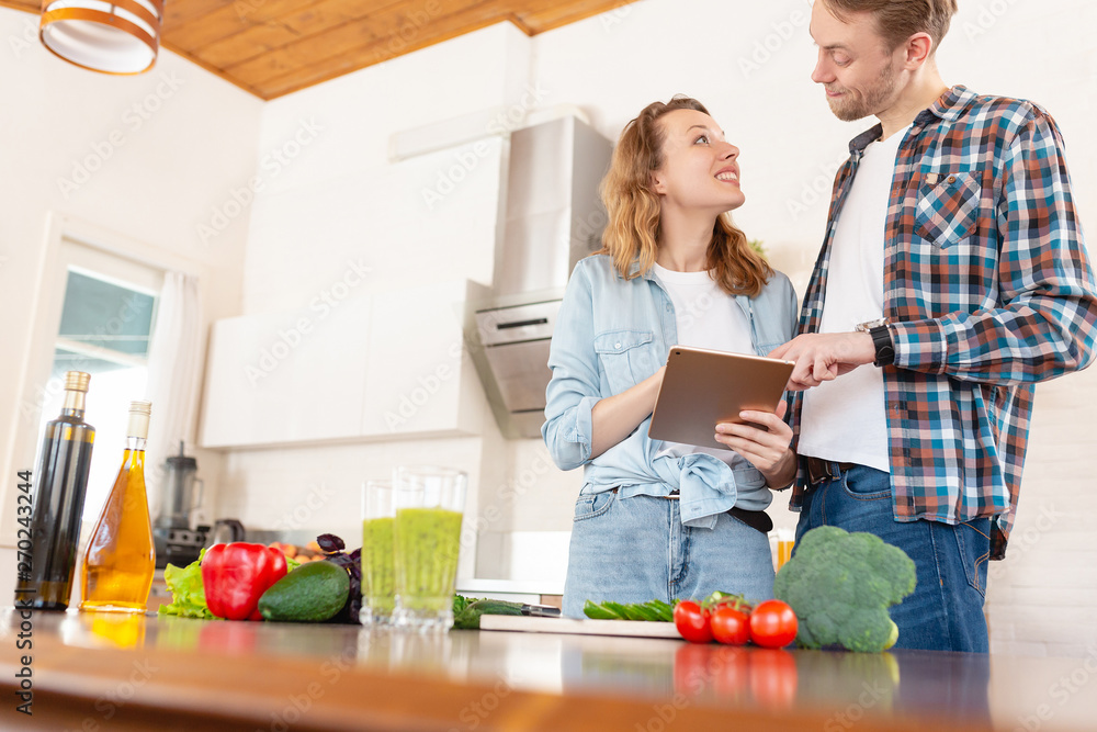 Young married couple in casual clothes is looking for a salad recipe that is made from vegetables using a smartphone and tablet while standing in the kitchen of their cozy country house Young married couple in casual clothes is looking for a salad recipe that is made from vegetables using a smartphone and tablet while standing in the kitchen of their cozy country house
