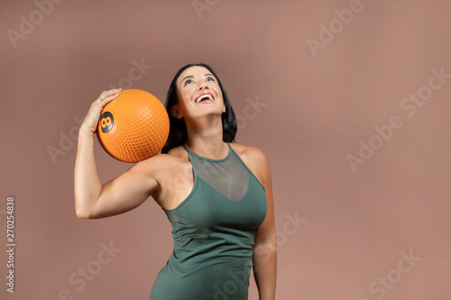 Beautiful brunette hair fitness model wearing white and green sportswear laughing holding an orange medicine ball on her shoulder against a brown background
