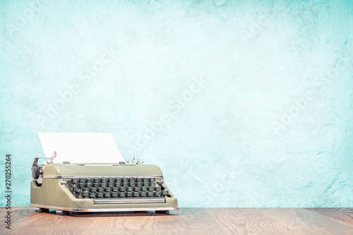 Retro classic typewriter from circa 50s with sheet of paper on wooden desk front textured aquamarine wall background. Vintage old style filtered photo