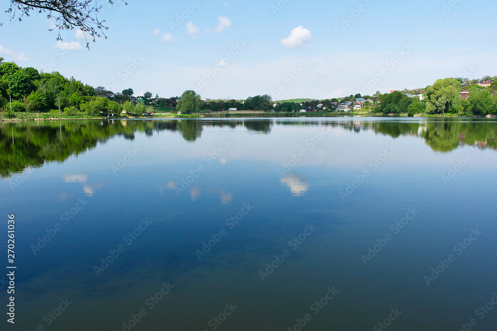 A lonely little cloud is reflected in a picturesque lake. Beautiful clean lake among the village.