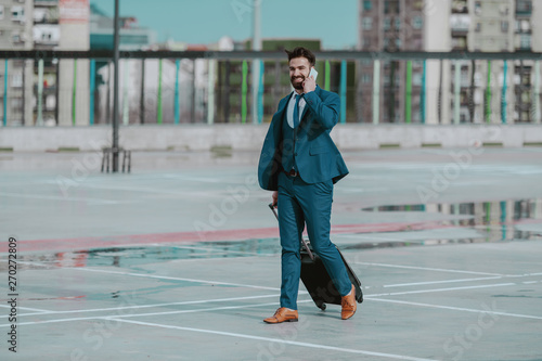 Young perspective smiling bearded Caucasian businessman in formal wear talking on the phone while carrying luggage and walking on the parking lot. Business trip concept.