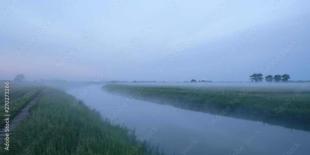 Trees on fog-covered green fields and meadows at a canal during sunrise in Havelland in Brandenburg, Germany 