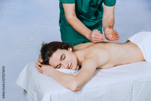 Massage therapist doing massotherapy of a young woman. Beautiful relaxed face of a young woman with brown hair and closed eyes on a blue background.