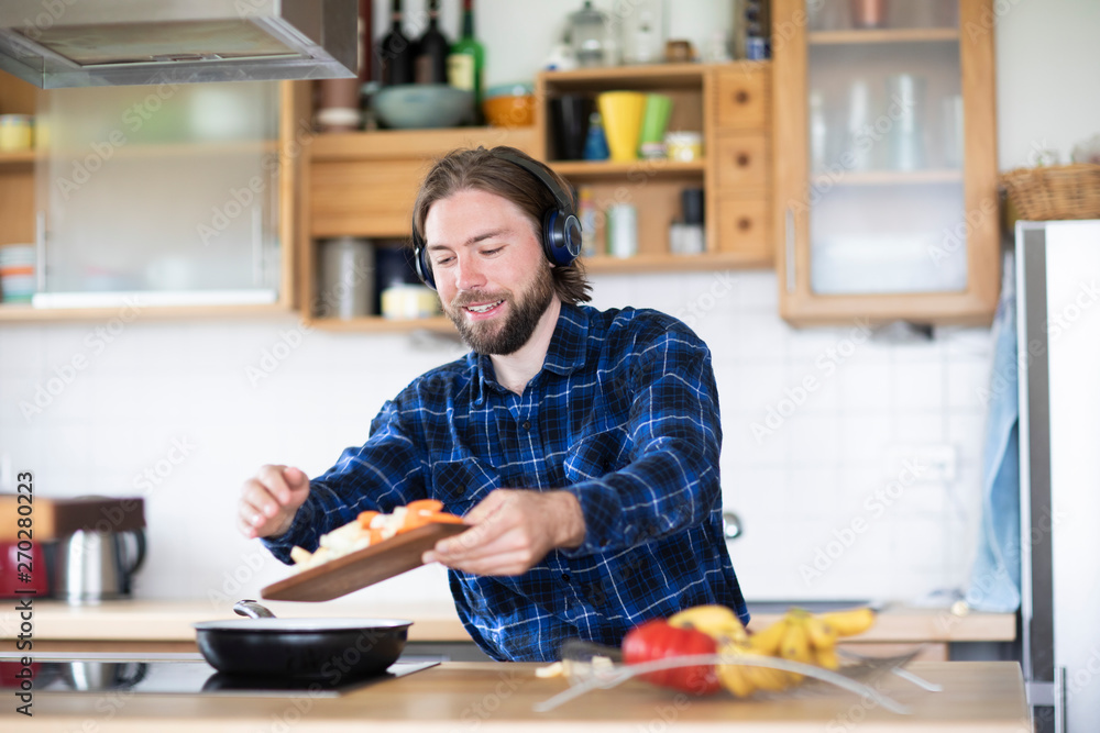 Young man with beard and plaid shirt, and headphones cooking vegetables in kitchen Young man with beard and plaid shirt, and headphones cooking vegetables in kitchen