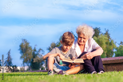 Attractive grandmother with her grandson are reading a book outdoors together. Summer family leisure. Self-education and development