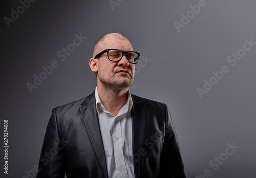 Thinking bald business man looking up serious in eyeglasses and have an idea in suit on grey background. Closeup