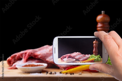 hands of man with smartphone taking photo Raw Ribs on a rustic cutting board with salt, pepper and grinder for spices . Black Background for copy space.