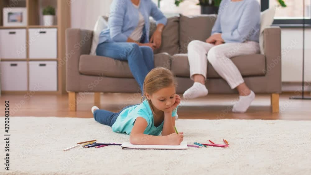 family, childhood and leisure concept - girl drawing in sketchbook by crayons lying on floor and adults sitting on sofa at home family, childhood and leisure concept - girl drawing in sketchbook by crayons lying on floor and adults sitting on sofa at home