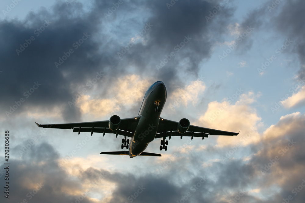 Obraz premium Travel, transportation, aviation concept. Silhouette of wide-body passenger airplane flying overhead after take off against a evening cloudy sky at sunset, bottom view