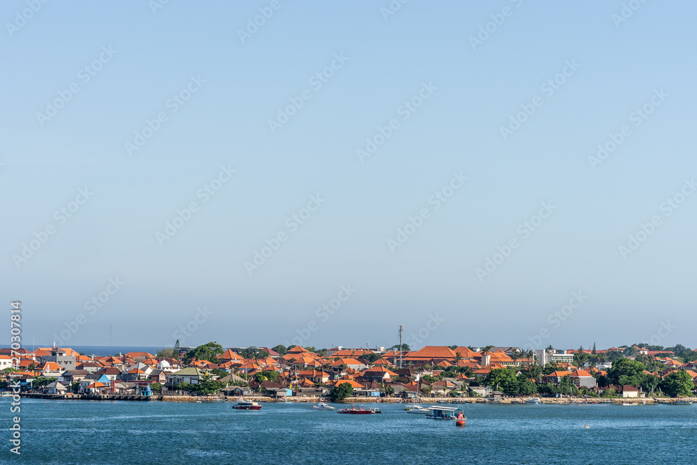 Benoa harbour, Bali, Indonesia - February 26, 2019: The red roofs of the buildings on Tanyung Benoa Peninsula under light blue sky, above dark blue water, seen from harbour. Green foliage, boats.