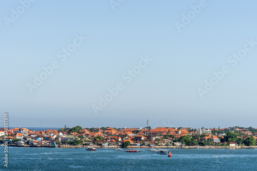 Benoa harbour, Bali, Indonesia - February 26, 2019: The red roofs of the buildings on Tanyung Benoa Peninsula under light blue sky, above dark blue water, seen from harbour. Green foliage, boats.