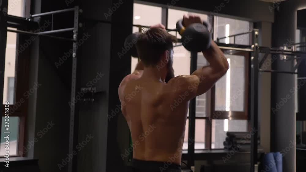 Closeup back view shoot of adult muscular athletic man making squats with the kettlebells indoors in the gym Closeup back view shoot of adult muscular athletic man making squats with the kettlebells indoors in the gym