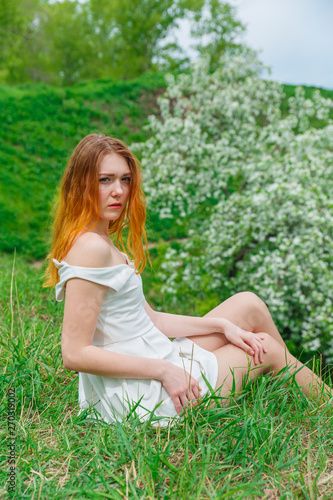 The red-haired girl in a white dress sitting on green grass on a background of a blossoming Apple tree. Girl spring on nature.
