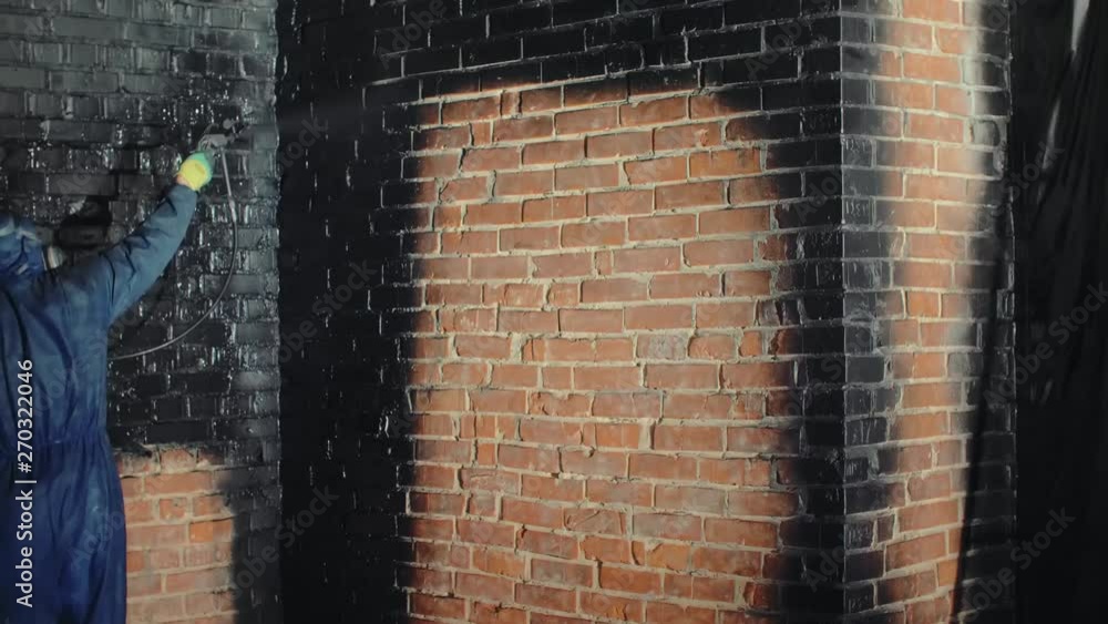 Worker wearing blue workwear painting the brick wall in black color by
