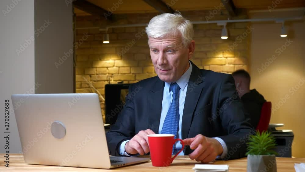 Closeup shoot of old caucasian office worker using the laptop and drinking coffee indoors in the office on the workplace Closeup shoot of old caucasian office worker using the laptop and drinking coffee indoors in the office on the workplace