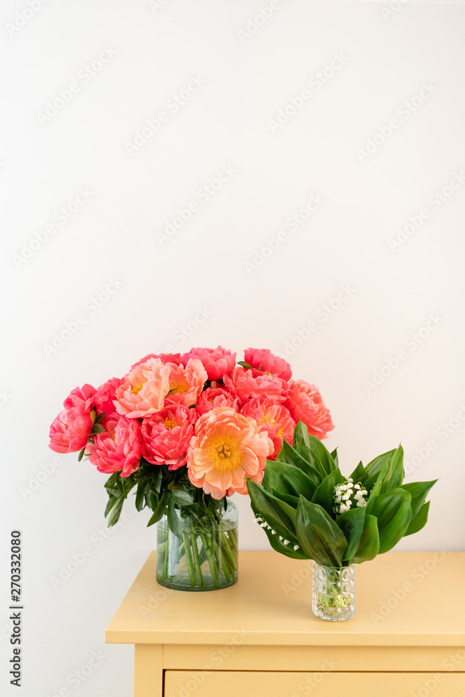 Fototapeta premium Coral peonies and spring lilies of the valley in a glass vase on wooden table.. Beautiful peony flower for catalog or online store. Floral shop concept . Beautiful fresh cut bouquet. Flowers delivery.