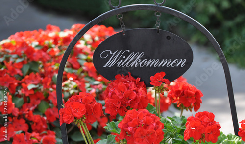 Close up of traditional austrian metal welcome sign with red flowers. German word Willkommen (welcome).