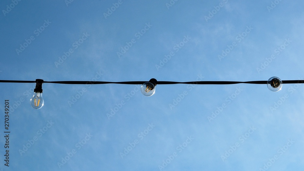 A string wire of outdoor light bulbs hanging against a clear blue sky. Light bulbs on the sky background. Panoramic view. A string wire of outdoor light bulbs hanging against a clear blue sky. Light bulbs on the sky background. Panoramic view.