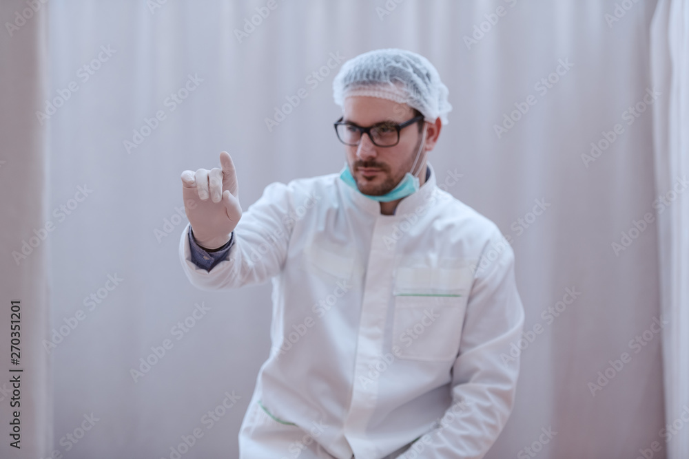 Young serious Caucasian male doctor in sterile white uniform and with eyeglasses pointing with finger.