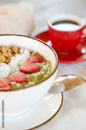 Delicious organic green smoothie bowl garnished with strawberries, sliced almonds and granola served with a cup of coffee