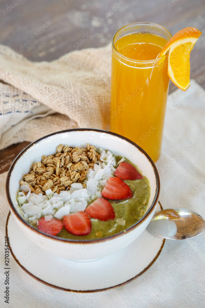 Delicious organic green smoothie bowl garnished with strawberries, sliced almonds and granola served with a glass of fresh squizzed orange juice