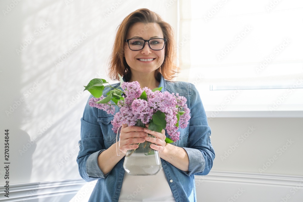 Portrait of beautiful mature woman at home with bouquet of lilac flowers. Background of home interior dining near the window Portrait of beautiful mature woman at home with bouquet of lilac flowers. Background of home interior dining near the window