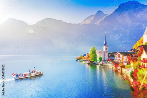 Colorful autumn seasonal scene of Hallstatter see lake in Austria and UNESCO historical village Hallstatt. Passenger boat glade by calm lake water, beautiful sunrise scenery.