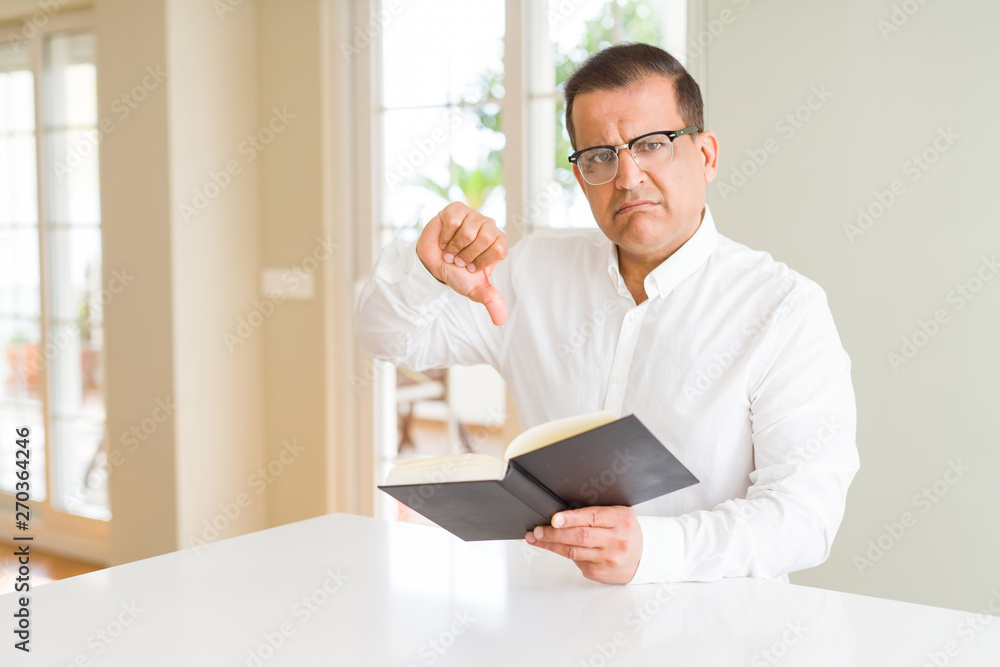 Middle age man reading a book at home wearing glasses with angry face, negative sign showing dislike with thumbs down, rejection concept