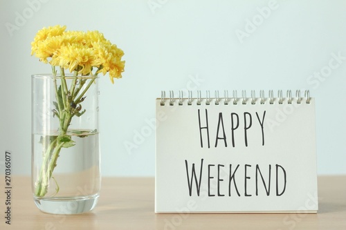 Yellow flower in glass with happy weekend notebook on wood table on white background. photo