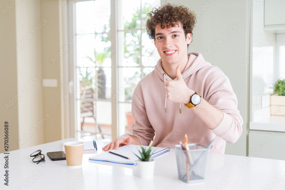 Young student man writing on notebook and studying doing happy thumbs up gesture with hand. Approving expression looking at the camera with showing success.