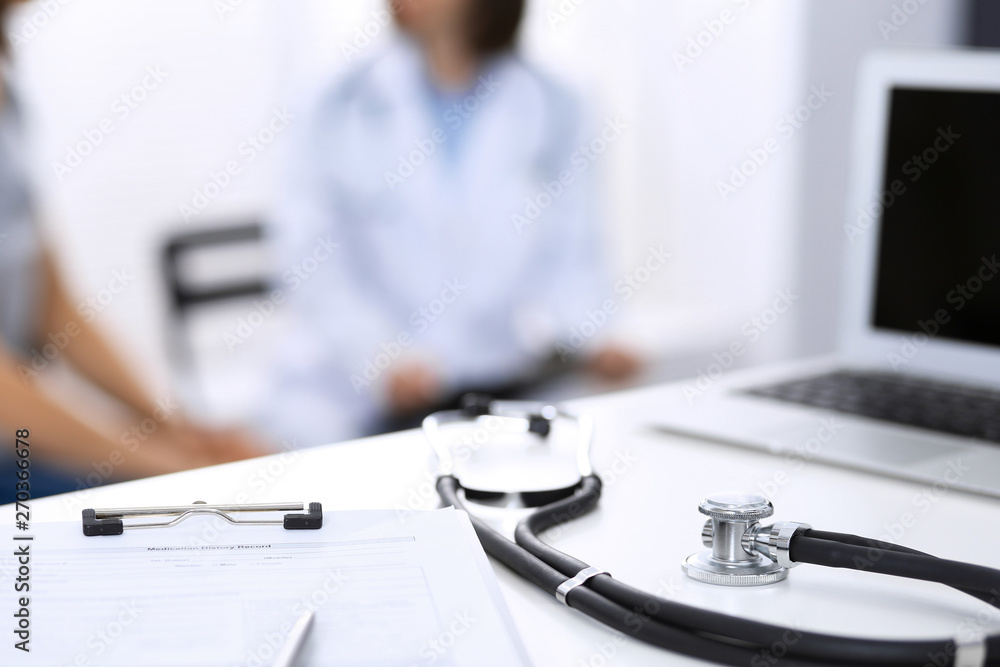 Stethoscope, clipboard with medical form lying on hospital reception desk with laptop computer and busy doctor and patient communicating at the background. Medical tools at doctor working table Stethoscope, clipboard with medical form lying on hospital reception desk with laptop computer and busy doctor and patient communicating at the background. Medical tools at doctor working table