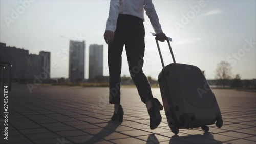 Close up, Knee or ground level shot: Business lady going to taxi parking from the airport or railroad station with her luggage in big sity. Slow motion. Girl in heels stepping and roll suitcase on