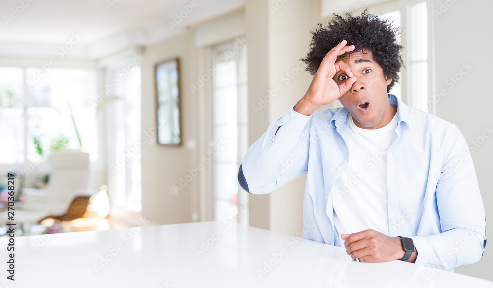 African American man at home doing ok gesture shocked with surprised face, eye looking through fingers. Unbelieving expression.