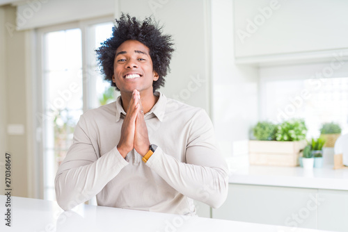 African American man at home praying with hands together asking for forgiveness smiling confident.