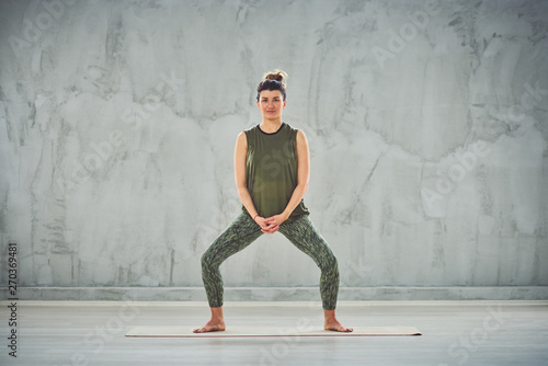 Full length of charming fit Caucasian brunette dressed in green outfit standing on mat and doing Goddess yoga pose.