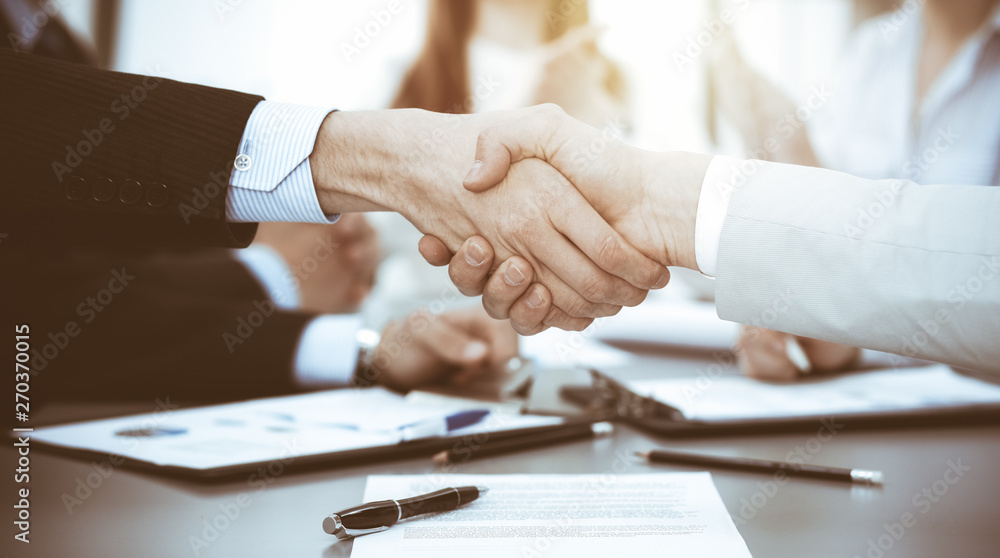 Business people shaking hands at meeting while theirs colleagues clapping and applauding. Group of unknown businessmen and women in modern white office. Success teamwork, partnership and handshake