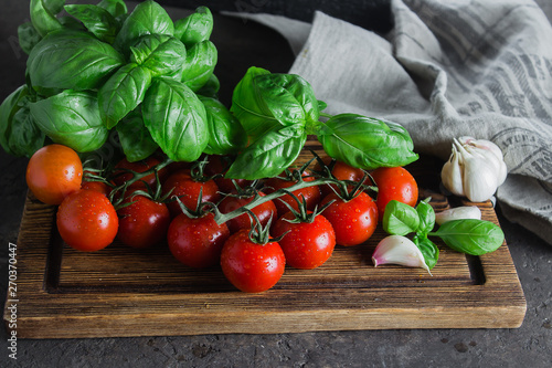 Organic Cherry Tomatoes with basil and garlic on cutting board on dark brown concrete stone background. Top view