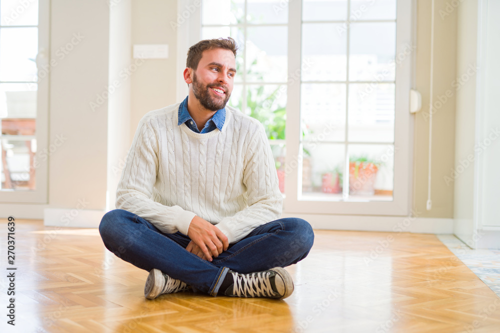 Handsome man wearing casual sweater sitting on the floor at home looking away to side with smile on face, natural expression. Laughing confident.