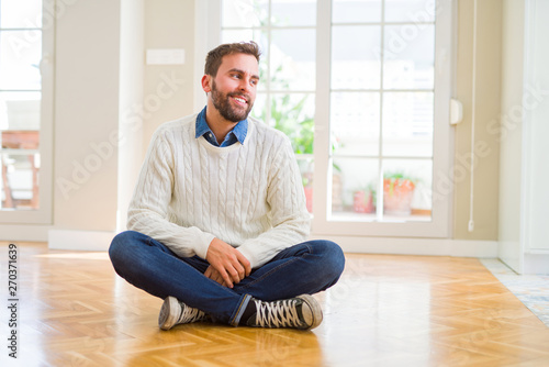 Handsome man wearing casual sweater sitting on the floor at home looking away to side with smile on face, natural expression. Laughing confident.